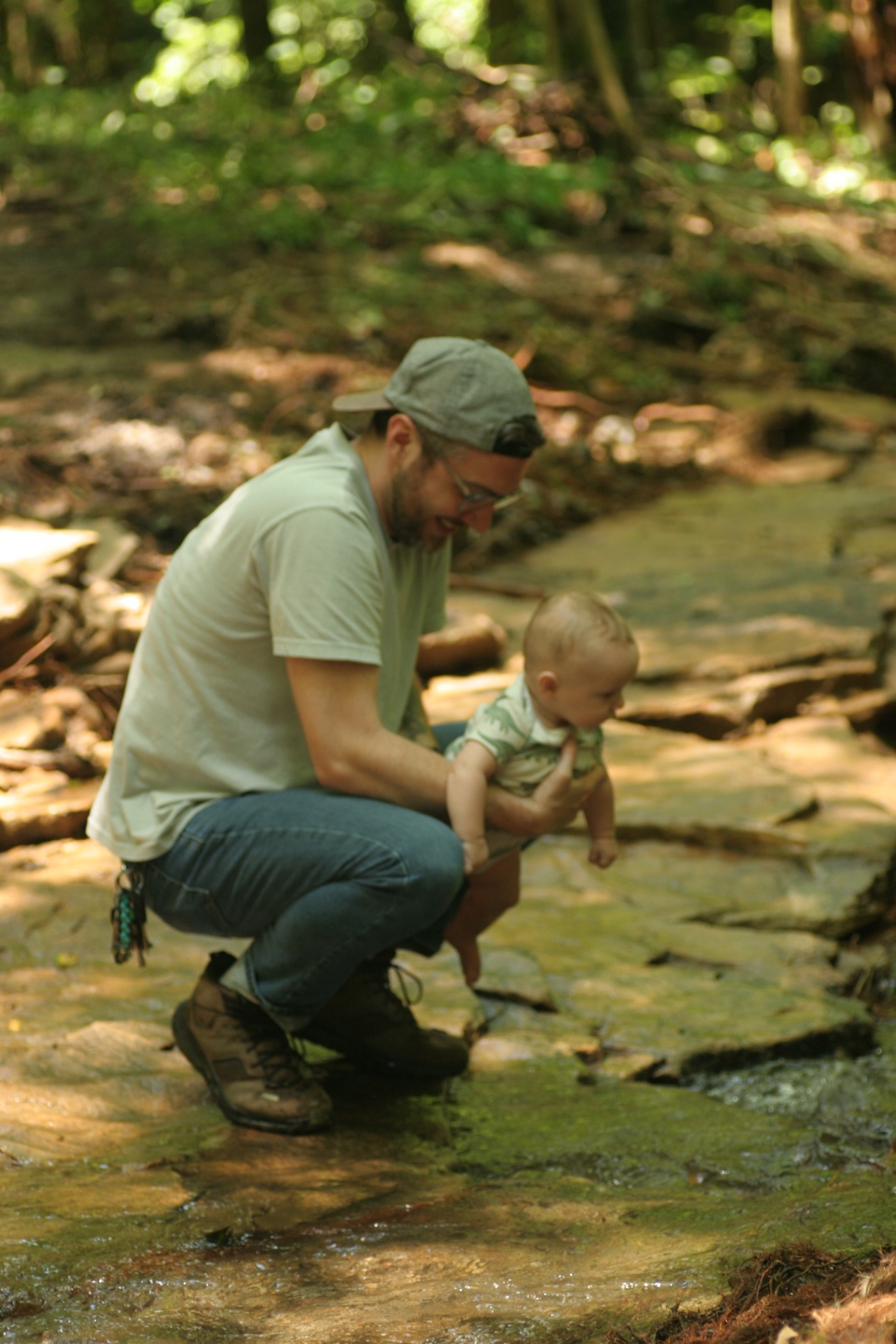 A father crouched on creek rock holding a baby toward the water