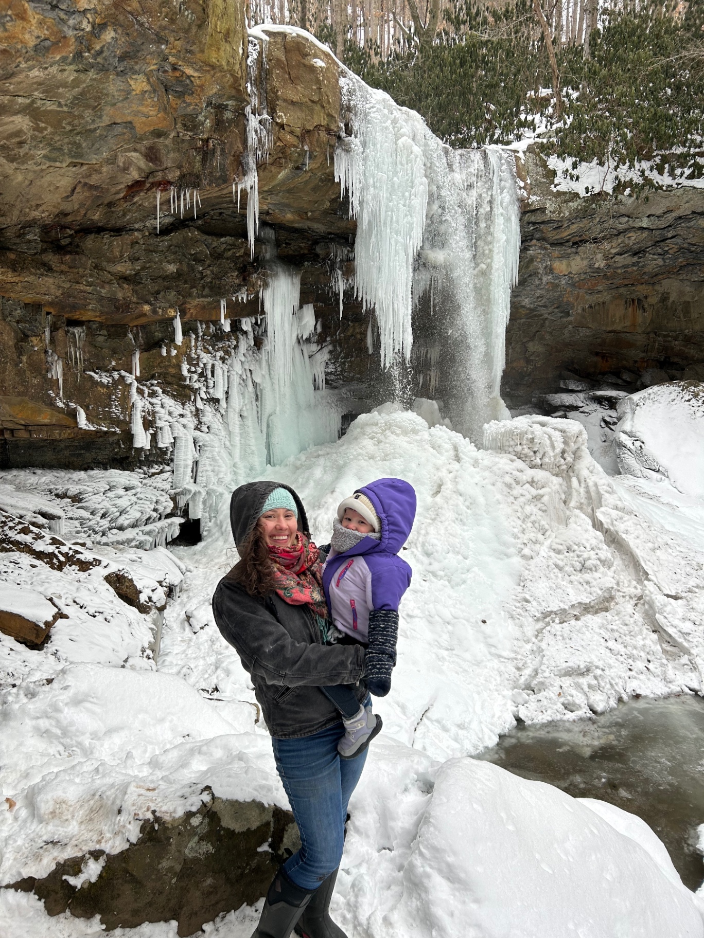 A parent holding a toddler in a purple snowsuit in front of a massive frozen waterfall